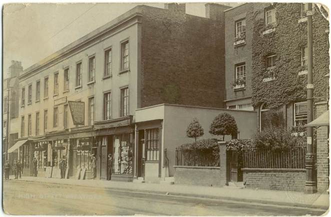 three-storey property block with shops, their owners standing in the doorways, then a three-storey property set back from the road