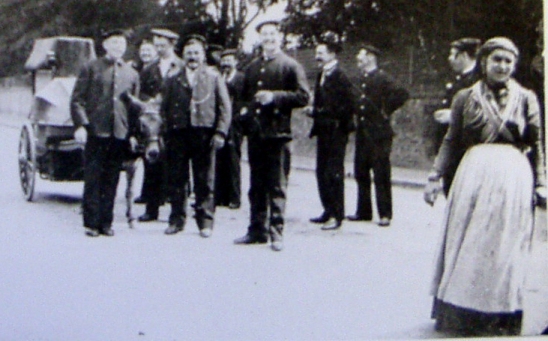 Group of 9 cheerful firemen leading a donkey and small organ; lady in traditional canal costume in foreground