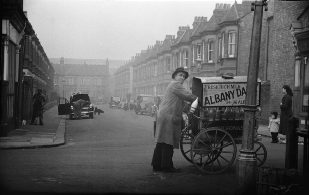 B/W photo of road, cars parked either side, milkman with hand cart in foreground