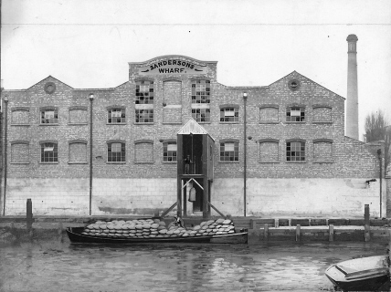 Originally sepia photo showing barge, heavily laden with sacks, beside 'Sandersons' wharf, a sack is being unloaded by a man standing on the gantry