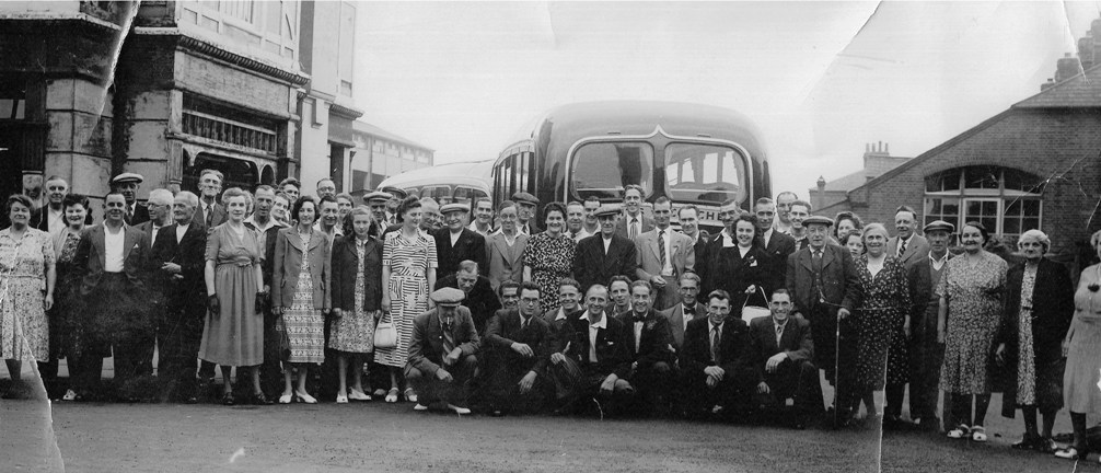 Crowd of over 50 people, range of ages but nobody under 12, ready to get on a coach (from Colchester?).  Ladies are dressed in print dresses, some with jackets or coats, some of the men have open-necked shirts, some wear ties.