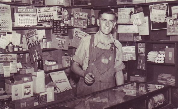 B/w photo of Stanley Newman at the counter, with an array of items pinned up behind him